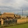 The image shows a secured military compound or motor pool viewed from outside a chain-link fence topped with barbed wire. Several tan/desert-colored military cargo trucks, consistent with LMTV or FMTV-type vehicles, are parked on a dirt surface inside the perimeter. A utility pole, a metal building, and additional vehicles are visible in the background.