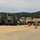 A military staging area or motor pool showing a row of military vehicles parked on a gravel surface. A single soldier in ACU (Army Combat Uniform) camouflage is walking toward the vehicles. The setting appears to be a training installation or base, surrounded by dense tree lines.