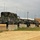 A row of military vehicles, primarily HMMWVs (Humvees) in woodland camouflage and tan paint schemes, are parked behind concrete Jersey barriers at what appears to be a military installation or staging area. The vehicles are equipped with multiple radio antennas, suggesting communications or command roles. The setting includes a dirt road, utility poles, and a white building structure in the background.