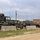 A row of military HMMWV (Humvee) vehicles is parked in an outdoor staging area, separated from a dirt road by concrete Jersey barriers. The vehicles display woodland camouflage paint schemes along with tan/desert variants, and are equipped with multiple radio antennas. The setting appears to be a military installation or training base, with utility poles and a white building visible in the background.