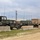 A military vehicle staging area or motor pool showing multiple HMMWV (Humvee) vehicles and at least one larger military truck parked in a gravel/dirt lot. The vehicles display a mix of woodland camouflage and tan/desert color schemes, with multiple radio antennas visible on each. Concrete Jersey barriers are positioned in the foreground, suggesting a security perimeter or access control point.