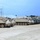 A row of M113 Armored Personnel Carriers (APCs) and associated military wheeled vehicles are parked in a gravel staging area at what appears to be a military installation or training base. The vehicles are painted in desert tan camouflage and several have canvas covers over their top hatches. The setting includes utility buildings and power lines in the background under a partly cloudy sky.