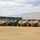 A row of military HMMWV (Humvee) vehicles is parked in a dirt lot at what appears to be a military installation or depot. The vehicles are predominantly desert tan in color, with at least two in olive drab/camouflage green. A large white metal warehouse building is visible in the background.