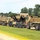 A convoy of U.S. Army tactical military vehicles is staged or moving along a road bordered by grass and trees. The vehicles are painted in desert tan/sand color and include heavy transport trucks and at least one HMMWV (Humvee) loaded on a flatbed carrier. Unit markings are visible on several vehicles, including '926 EN BDE' and '841 EN BX'.
