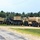 A row of military vehicles in desert tan/beige camouflage paint is parked along a gravel area beside a road, with a tree line visible in the background. The vehicles include heavy tactical trucks and fuel tankers marked with 'FLAMMABLE' hazard labels. The setting appears to be a military installation or training base during summer conditions.