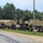 A row of military tactical vehicles is parked along a gravel area beside a paved road, with a grassy strip and dense tree line in the background. The vehicles appear to be US Army heavy transport trucks in desert tan/sand camouflage paint. The setting appears to be a military installation or training base.