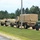 A convoy of U.S. military vehicles is parked in a line along a gravel area adjacent to a grassy field, likely at a military installation or training base. The vehicles include multiple HMMWVs (Humvees) and at least one larger tactical truck with a canvas cover, along with a green water/fuel trailer. The setting appears to be a summer day with dense tree coverage in the background.
