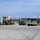 A military vehicle staging area showing a civilian semi-truck flatbed transporter carrying a tan military tactical truck, alongside a row of tan HMMWVs (Humvees) lined up in a paved lot. The scene appears to be at a National Guard or military installation, with warehouse-style buildings and a shipping container visible in the background. A civilian worker is visible near the flatbed truck, suggesting active loading or unloading operations.