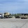 A military staging area or motor pool showing a civilian semi-truck with a flatbed trailer transporting a tan military tactical truck (likely an LMTV or FMTV). Several tan military Humvees are lined up on the right side of the paved lot, with a few civilian vehicles and personnel visible in the background. The scene appears to be a military installation with industrial buildings and a shipping container visible.