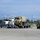A civilian flatbed semi-truck is transporting a military tactical vehicle (appears to be an LMTV or similar tan/desert-colored Army truck) in what appears to be a military installation or staging area. Two civilians and at least one uniformed military personnel are visible near the vehicles. Additional military Humvees and shipping containers are visible in the background.