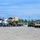 A military vehicle staging area showing a civilian semi-truck (blue and white Kenworth) with a flatbed trailer carrying a tan military tactical truck, likely an LMTV or FMTV. Several Humvees (HMMWVs) are lined up in the background alongside a green shipping container. The scene appears to be a military installation or National Guard armory with personnel overseeing equipment loading or transport operations.