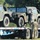 Two military personnel in camouflage uniforms are loading or securing HMMWV (Humvee) military vehicles onto a flatbed trailer. One soldier is standing on the trailer bed near the vehicle door, while a second soldier stands on the ground below. The scene appears to be a vehicle transport or deployment operation at a military installation.