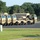 A formation of tan/desert-camouflage military vehicles is lined up in a paved lot, likely on a military installation. The vehicles include multiple large tactical trucks (appearing to be LMTV or FMTV variants) and several HMMWVs (Humvees), with at least one covered cargo truck visible. One or two soldiers in uniform are visible near the vehicles, suggesting pre-deployment staging or motor pool activity.