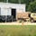 A military installation maintenance facility is shown with a tan/desert-camouflage tactical truck (likely an LMTV or FMTV) parked outside a white metal maintenance bay building. A woodland-camouflage painted trailer or equipment unit is positioned near the open bay doors. The setting appears to be a U.S. Army post or National Guard facility surrounded by trees.