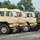 A row of military tactical trucks is parked in a paved lot, predominantly painted in desert tan (sand) camouflage with one vehicle in woodland green camouflage pattern. The vehicles appear to be LMTV or FMTV (Family of Medium Tactical Vehicles) style trucks, along with at least one HMMWV (Humvee) visible on the left. The setting appears to be a military staging area or motor pool during summer, indicated by the lush green trees in the background.