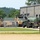 A military installation or motor pool area showing several military vehicles staged near a large maintenance facility building. The scene includes a mix of tan/desert-camouflage and olive drab military trucks, with a fuel or water tanker trailer prominently visible in the foreground. The area is secured by a chain-link fence and features a paved road with a posted speed limit sign.