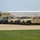 A large formation of military HMMWV (Humvee) vehicles is parked in a diagonal row on a gravel or dirt lot, alongside at least one larger military truck. The vehicles are painted in desert tan/sand coloring and are equipped with radio antenna mounts. A white metal warehouse or hangar building is visible in the background.