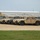 A large formation of military HMMWV (Humvee) vehicles is parked in a row on a gravel lot at what appears to be a military installation or motor pool. The vehicles are a mix of tan/desert and olive drab color schemes, all equipped with radio antennas. A large white metal warehouse-style building is visible in the background.