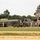 The image shows a military installation or motor pool area with several military vehicles parked behind a chain-link fence. A large green tanker truck and additional tactical military vehicles are visible, along with at least one uniformed soldier. The setting appears to be a U.S. Army base or National Guard facility during summer conditions.