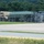 A military compound or motor pool area secured by a chain-link fence, containing multiple military tactical vehicles including what appears to be fuel tanker trucks in olive drab and tan camouflage colors. At least one uniformed military personnel is visible near the fence gate. The scene appears to be at a military installation or training area, with wooded hills visible in the background.
