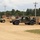 A military motor pool area showing multiple HMMWV (Humvee) vehicles parked on a gravel/dirt surface. At least one soldier in camouflage uniform is visible near the vehicles. The setting appears to be a training base or staging area, with shipping containers and utility poles visible in the background.