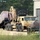 A tan/desert-camouflage military heavy-lift vehicle, consistent with a US Army Palletized Load System (PLS) truck, is maneuvering near a warehouse building. The vehicle appears to be loading or unloading palletized cargo wrapped in plastic shrink-wrap onto a flatbed trailer. The scene is set on a gravel/dirt surface at what appears to be a military installation or logistics depot.