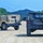 Two military HMMWV (Humvee) vehicles are traveling along a gravel/dirt road in a training area. The vehicle in the foreground is painted in woodland camouflage pattern while the vehicle in the background is painted in desert tan. Both vehicles are equipped with radio antennas and soft-top canvas enclosures.