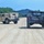 Two military HMMWV (Humvee) vehicles are traveling along a gravel/dirt road in a training area surrounded by trees and hills. The vehicle on the right is painted in woodland camouflage pattern, while the vehicle on the left is painted in desert tan. Both vehicles are equipped with radio antennas and appear to be part of a convoy or patrol exercise.
