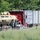 The image shows a staging or transport area where a tan/desert-colored military armored wheeled vehicle (likely an M1117 Guardian or similar MRAP-type vehicle) is positioned on a lowboy flatbed trailer. A red semi-truck cab is hauling a white intermodal shipping container marked 'USAU 092010' nearby. At least one person is visible between the vehicles, and the scene appears to be in a rural or semi-wooded area.