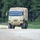 A tan/sand-colored military tactical truck, consistent with an LMTV (Light Medium Tactical Vehicle) or FMTV variant, is traveling along a paved road in a wooded area. The vehicle features a canvas-covered cargo bed and large off-road tires, indicating military utility transport use. The setting appears to be a military installation or training area based on the road type and surrounding terrain.