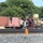 The image shows a convoy of semi-trucks transporting military equipment and shipping containers along a road adjacent to a railroad track. At least one truck is carrying what appears to be military vehicles or heavy equipment in desert tan/camouflage coloring, while others haul red intermodal shipping containers. A railroad crossing marker with an orange and red diagonal sign is visible in the foreground.