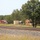 The image shows a rural area alongside a railroad track with multiple vehicles parked or staged on a gravel lot. A mix of military and civilian trucks are visible, including what appears to be a military utility truck, a red semi-truck, and an orange/yellow semi-truck cab. The scene is set against a backdrop of dense green trees under a clear sky.