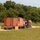 A semi-truck with a dark red/maroon cab is hauling a flatbed trailer loaded with a large orange intermodal shipping container on a dirt/gravel road. The scene is set in a rural, open field environment bordered by dense green trees. Additional equipment or machinery appears to be loaded behind the cab alongside the container.