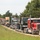 A convoy of civilian flatbed semi-trucks is transporting military vehicles along a rural road lined with trees and grass. The lead truck is a red Peterbilt hauling what appears to be a tan/beige MRAP or similar armored military vehicle. Multiple trucks stretch back along the road, carrying additional military equipment including what appears to be a camouflage-painted military truck on one of the flatbeds.