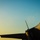 A close-up silhouette photograph of a military fighter jet, likely an F-35 or similar modern stealth aircraft, captured during golden hour lighting conditions. The image shows the forward fuselage, cockpit canopy, wing, and vertical stabilizer against a gradient blue-to-yellow sky. Warm sunset light illuminates portions of the aircraft's upper surfaces, revealing some surface details and a small insignia.