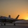 A fifth-generation stealth fighter jet, identifiable as an F-35 Lightning II, is parked on a tarmac at sunset. The aircraft bears a roundel insignia consistent with the Royal Australian Air Force (RAAF) on its fuselage. The dramatic golden-hour lighting silhouettes the aircraft against a mountain range backdrop, with a bird visible in flight in the upper right.