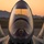 A military fighter jet, consistent with an F-22 Raptor or F-35, is photographed head-on from the front on an airfield tarmac during golden hour or sunset. A helmeted pilot is visible seated in the cockpit beneath the large, reflective canopy. The warm orange sky and distant mountain silhouette create a dramatic backdrop.