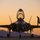 A fifth-generation stealth fighter jet, identifiable as an F-35 Lightning II, is photographed head-on during sunset or sunrise on a military airfield tarmac. The canopy is open and a ground crew member is visible walking on the aircraft's wing, likely conducting a pre- or post-flight inspection. The warm orange and purple sky with silhouetted mountains in the background creates a dramatic, high-contrast composition.