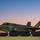 A fifth-generation stealth fighter jet, identifiable as an F-35A Lightning II, is parked on a tarmac at dusk or dawn. The aircraft bears Royal Australian Air Force (RAAF) markings, including the distinctive RAAF roundel and tail number '03'. The dramatic sunset/sunrise sky with silhouetted mountains provides a striking backdrop.