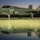 A fifth-generation stealth fighter jet, identified as an F-35A Lightning II bearing Australian Royal Australian Air Force (RAAF) markings, is parked on a tarmac at dusk or dawn. The aircraft displays the number '003' and features the RAAF roundel insignia on its fuselage. The wet tarmac surface creates a vivid mirror-like reflection of the aircraft beneath it, with a colorful orange and purple twilight sky in the background.