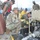 Military personnel from multiple branches, including U.S. Army soldiers in OCP camouflage and U.S. Marines in MARPAT uniforms, are gathered around a piece of military equipment, likely a generator or pump unit. The group appears to be conducting a training exercise, equipment inspection, or maintenance operation at what appears to be a military installation. Civilian or contractor personnel in yellow shirts are also present and actively involved.
