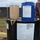 The image shows industrial fuel storage drums at what appears to be a military or aviation facility. The large black drum is labeled as 'Fuel System Icing Inhibitor, High Flash' (MIL-DTL-85470B), with smaller containers — one tan and one blue — placed on top. Personnel in military-style clothing are visible in the background near a fenced perimeter.