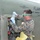 A U.S. Marine in camouflage uniform is performing maintenance or sampling work on a military fuel vehicle or tanker, identified by markings as flammable. The service member is wearing appropriate personal protective equipment including safety goggles, blue nitrile gloves, and a yellow apron. The work appears to involve collecting a fluid sample or servicing a fuel system component.