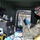A U.S. Marine in digital camouflage uniform is conducting water quality testing inside a mobile field laboratory unit. The Marine is wearing protective gear including safety goggles and blue nitrile gloves, along with a yellow apron, while handling liquid samples and testing equipment. In the background, other military personnel and vehicles are visible on what appears to be a gravel training or operational area.