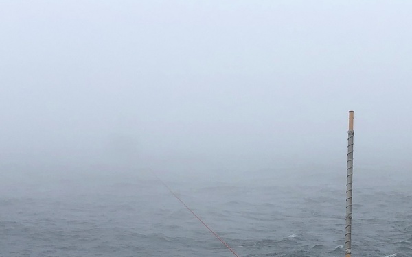 The image shows the bow deck of a vessel operating in rough, foggy sea conditions. A bright orange/red line extends from the ship into the churning water, suggesting an active towing, mooring, or scientific deployment operation. Visibility is severely reduced by dense fog, and the sea surface shows whitecaps indicating significant wave activity.
