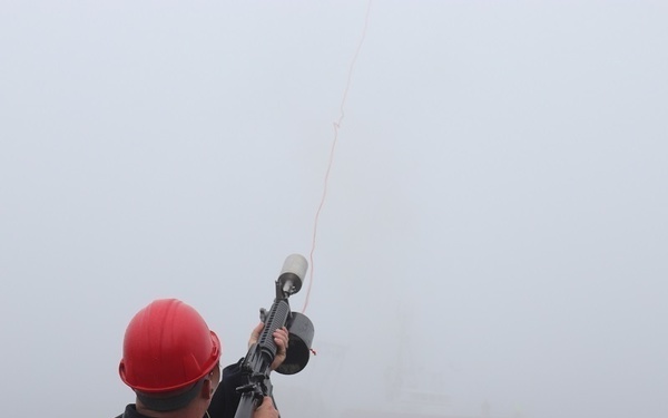A U.S. Coast Guard member is firing a line-throwing gun from the deck of a vessel in heavy fog conditions. The projectile and attached line are visible in the air, indicating a line-throwing or rescue line deployment exercise or operation. A second vessel is faintly visible in the foggy background.
