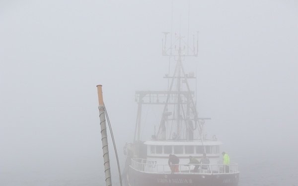 U.S. Coast Guard personnel aboard a cutter are conducting a boarding or assistance operation on a red commercial fishing vessel in heavy fog conditions. Multiple Coast Guard crew members wearing red life vests and hard hats are positioned on the deck, managing lines and preparing for vessel contact. The fishing vessel, partially obscured by dense fog, has several crew members visible on its deck.
