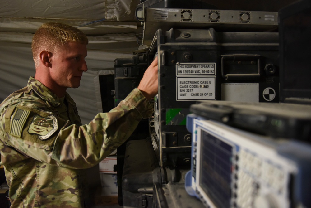 A U.S. Air Force service member in OCP (Operational Camouflage Pattern) uniform is working with military electronic equipment inside what appears to be a tent or temporary shelter. The equipment includes rack-mounted military-grade electronic cases with visible labels indicating operation at 120/240 VAC, 50-60 Hz. A test and measurement device, likely a spectrum analyzer or signal analyzer, is visible in the foreground.