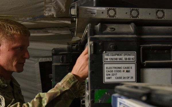 A U.S. Air Force service member in OCP (Operational Camouflage Pattern) uniform is working with military electronic equipment inside what appears to be a tent or temporary shelter. The equipment includes rack-mounted military-grade electronic cases with visible labels indicating operation at 120/240 VAC, 50-60 Hz. A test and measurement device, likely a spectrum analyzer or signal analyzer, is visible in the foreground.