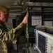 A U.S. Air Force service member in OCP (Operational Camouflage Pattern) uniform is working with military electronic equipment inside what appears to be a tent or temporary shelter. The equipment includes rack-mounted military-grade electronic cases with visible labels indicating operation at 120/240 VAC, 50-60 Hz. A test and measurement device, likely a spectrum analyzer or signal analyzer, is visible in the foreground.